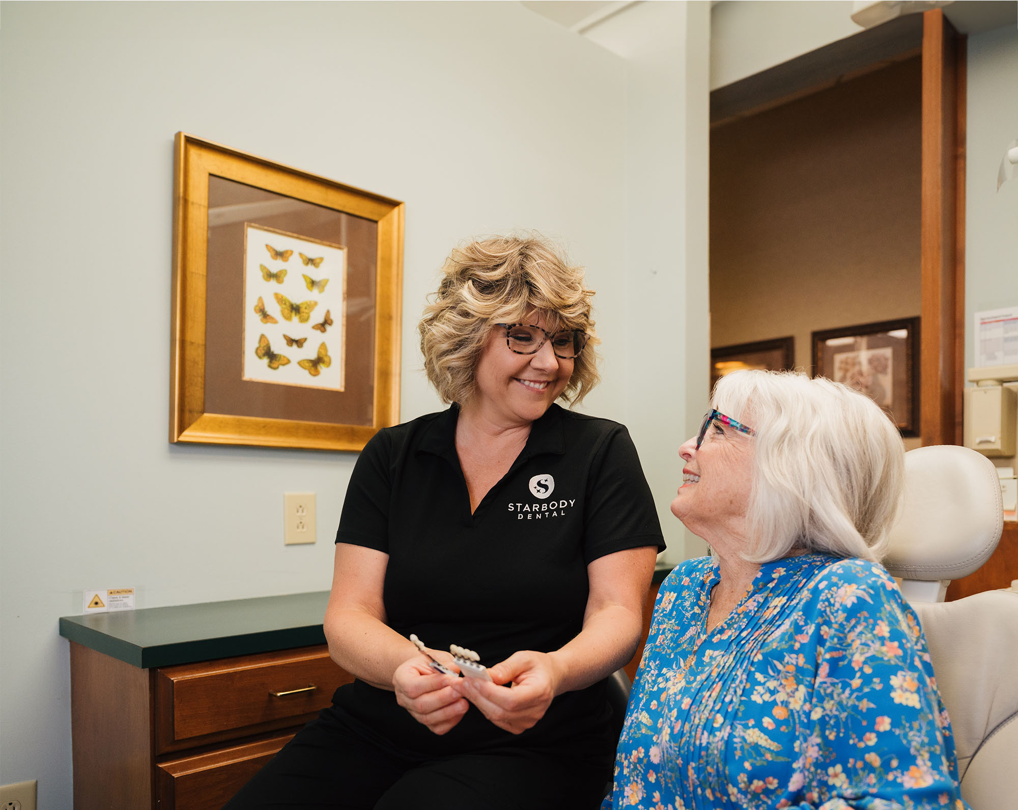 Dental professional in black Starbody Dental shirt showing a dental shade guide to an older woman in a blue floral blouse in a dental office.