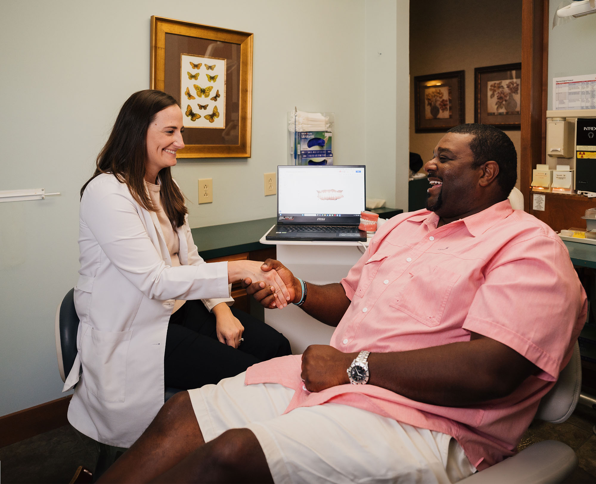 Smiling dentist in white coat shaking hands with a male patient sitting in a dental chair.