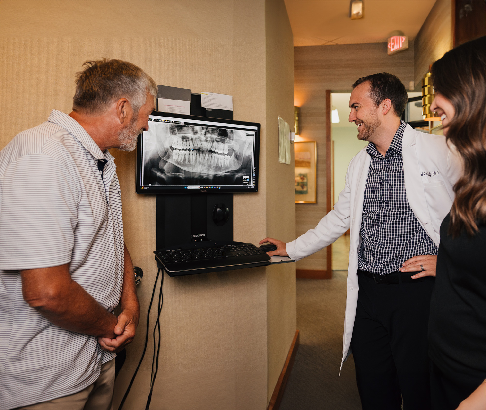 Dentist in a white coat showing a dental X-ray on a monitor to a smiling older male patient in a dental office.