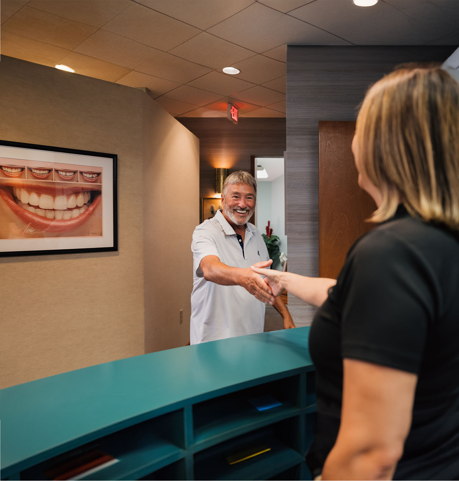 Smiling man in a white polo shirt shaking hands with a woman across a teal reception desk in a dental office.