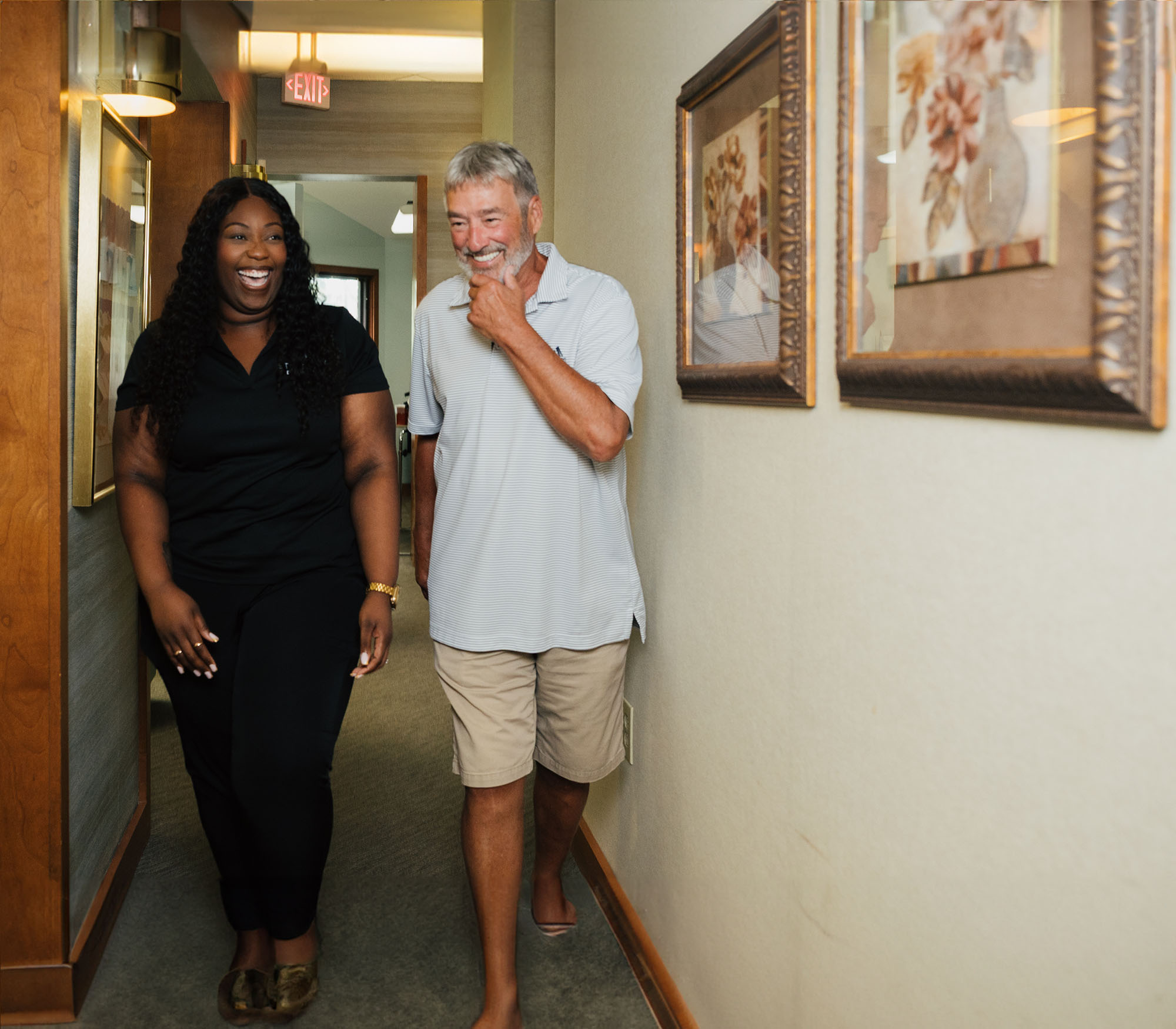 Smiling woman in black outfit walking down a hallway with a barefoot man in a gray shirt and beige shorts.