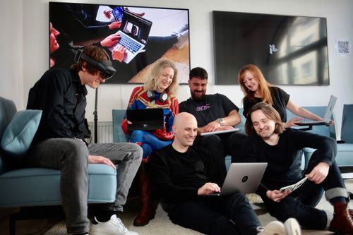 Eine Gruppe von Personen sitzt teilweise auf dem Sofa und auf dem Boden und schauen zusammen in ein Laptop. Auf diesem findet eine Online-Konferenz statt.