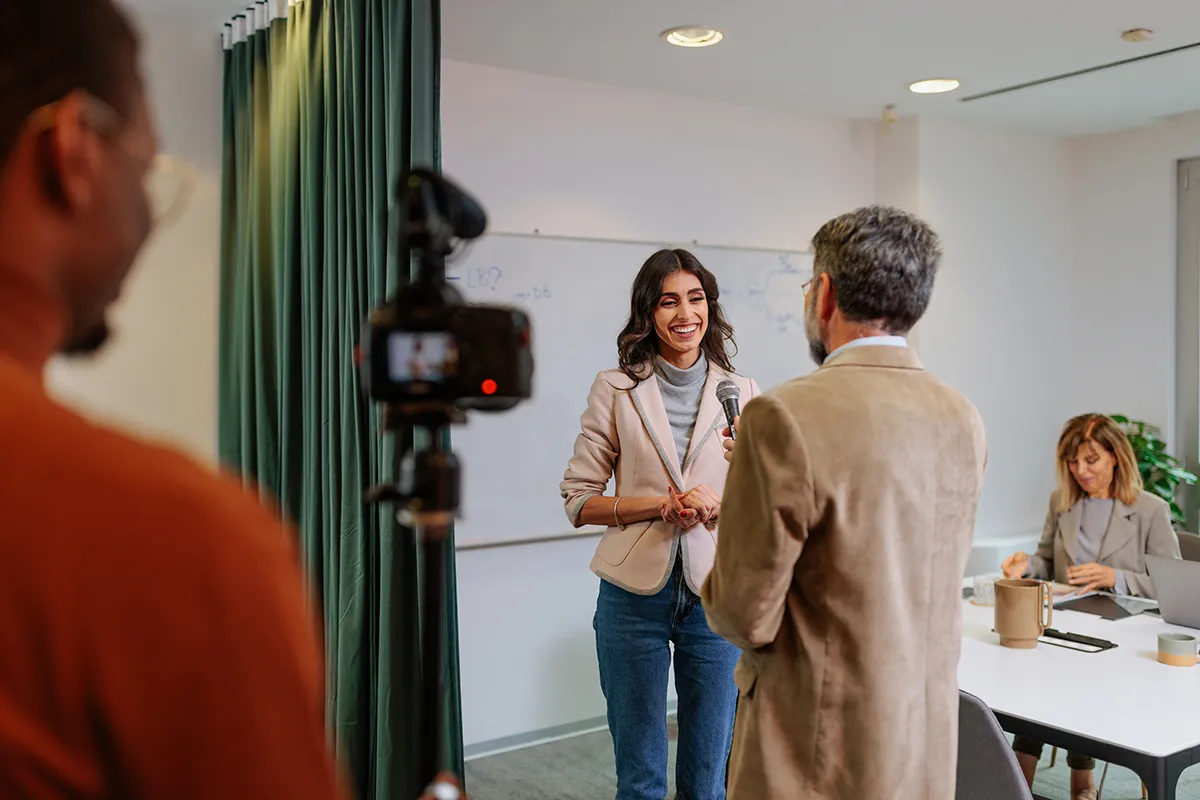 A woman speaking into a microphone during an interview in a conference room, with a camera filming in the foreground and two colleagues working at a table in the background.