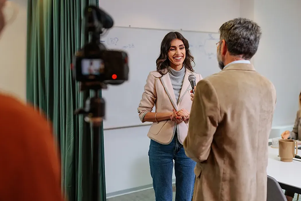 A woman speaking into a microphone during an interview in a conference room, with a camera filming in the foreground and two colleagues working at a table in the background.