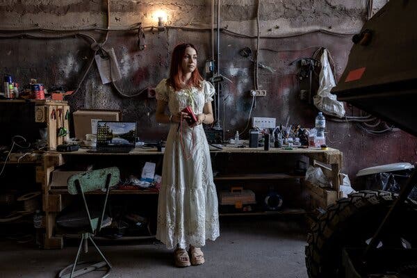 A young woman with red hair wearing sandals and a long white dress stands in front of a gritty cement wall and a work bench cluttered with tools, spare parts and assorted tech gear.