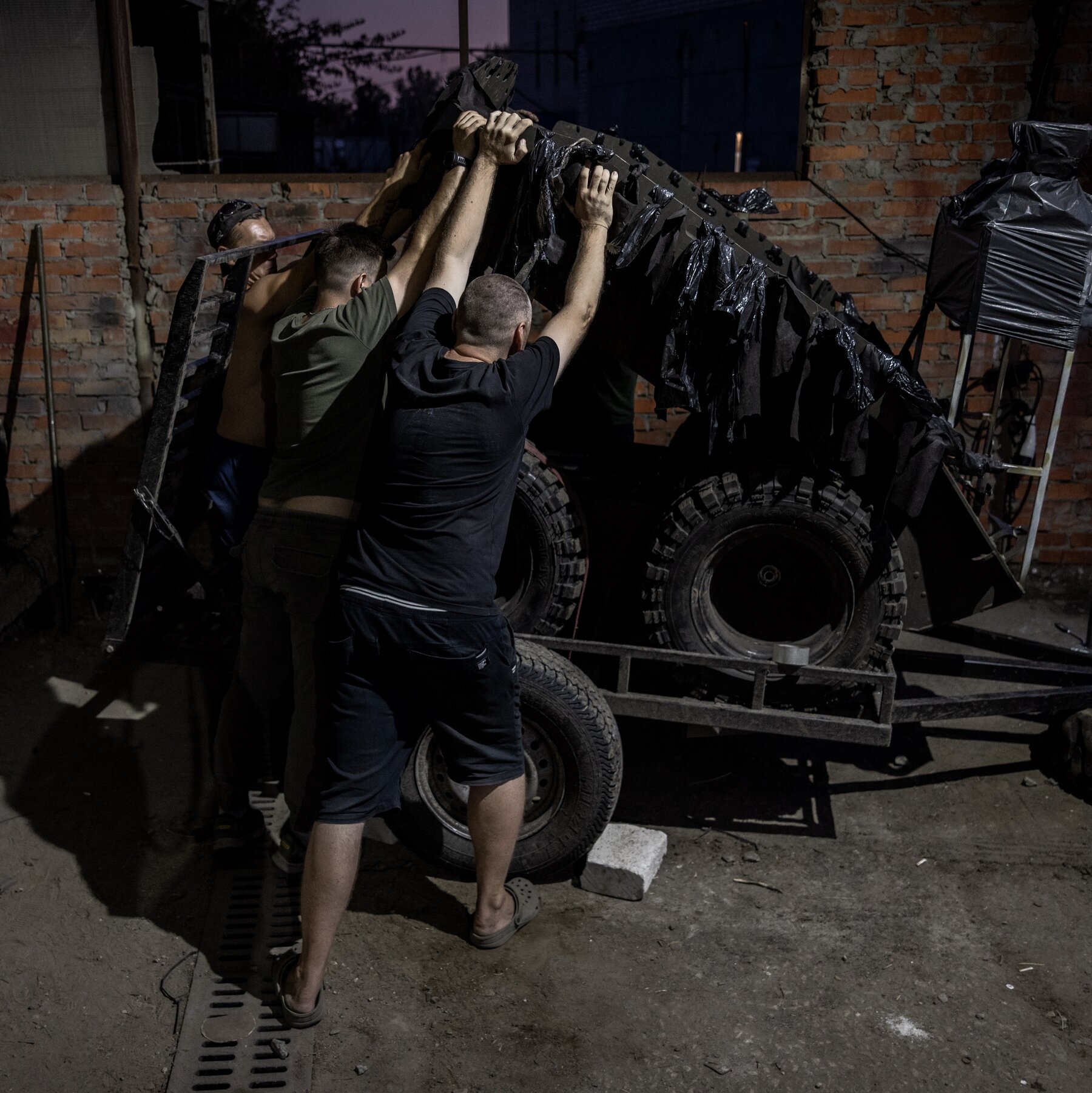 Three men wearing T-shirts, their faces obscured from the camera, lift a ground drone off a small trailer in a gritty brick warehouse.