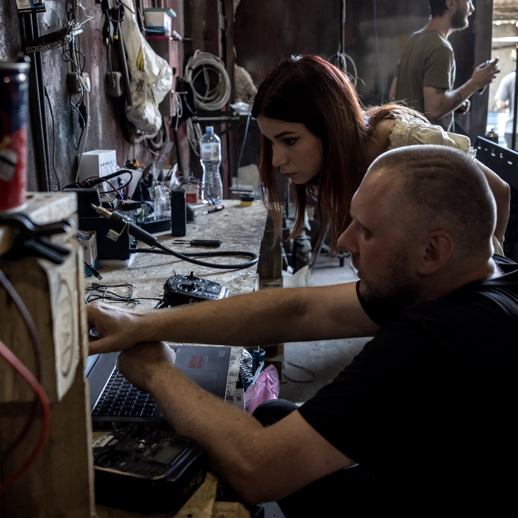 A man in a black T-shirt shows a young woman with red hair and a white dress something on a laptop as they sit at a workbench in a garage cluttered with spare parts and other mechanical gear.