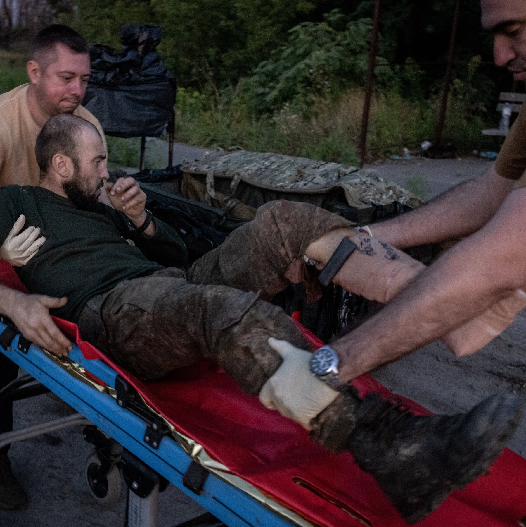 Two military medics in khaki tees lift a soldier with a wounded left leg and camouflage pants onto a stretcher in a wooded area.
