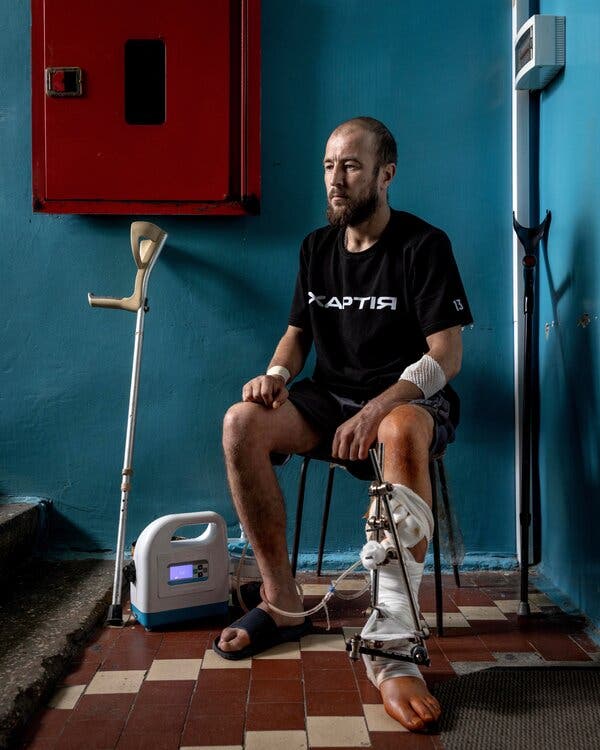 A man in a T-shirt and shorts, with bandages on his arms and a brace on his swollen left foot, sits in front of a blue wall with a single walking crutch standing next to him.