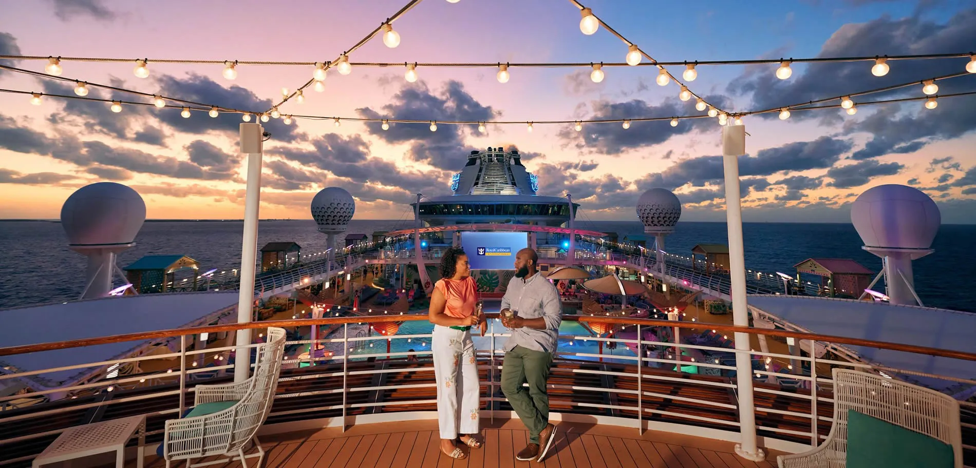Two people on cruise ship deck at sunset with string lights and ocean view