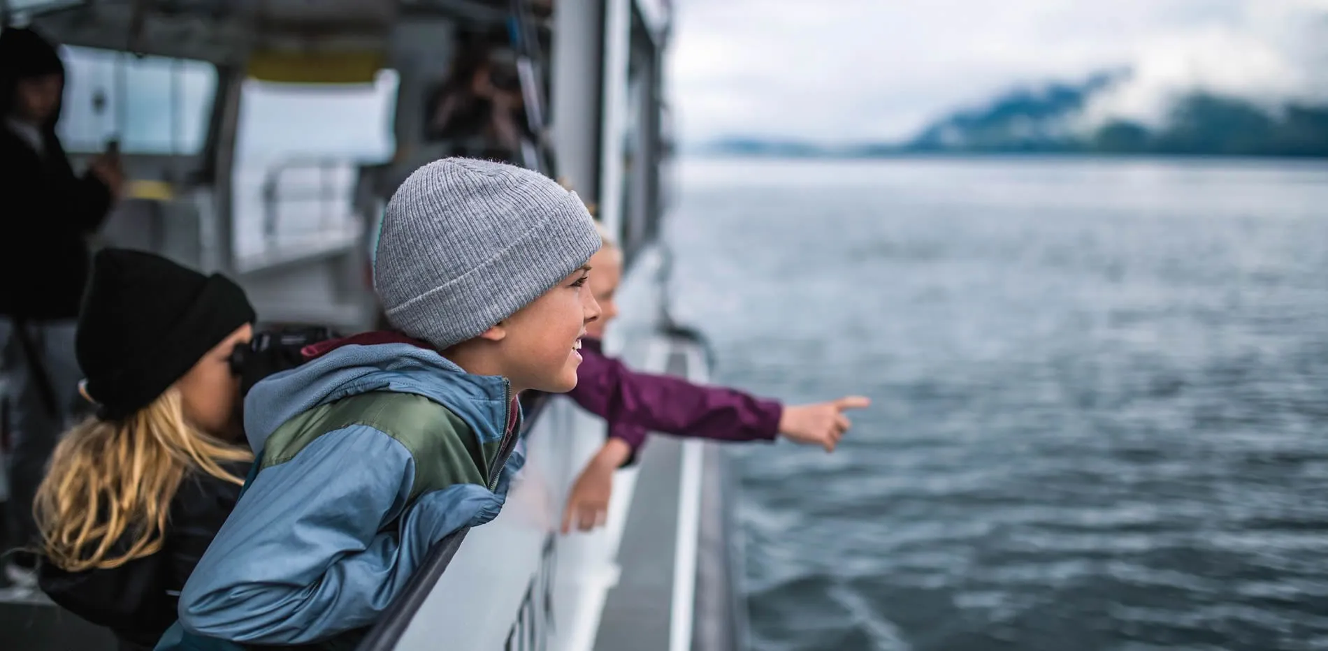 Child in gray beanie pointing excitedly from ferry with misty mountains behind