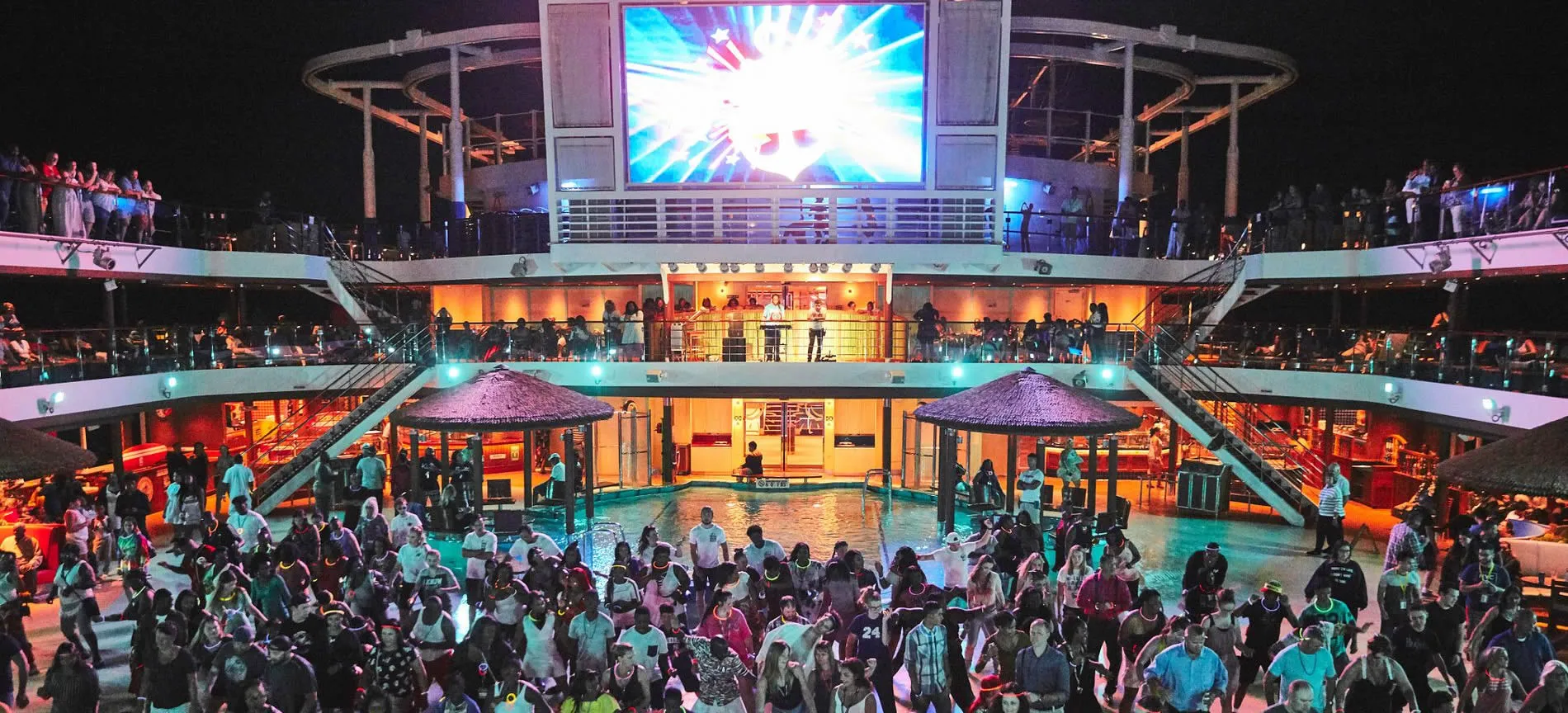 Crowded cruise ship deck at night with large screen and colorful lights