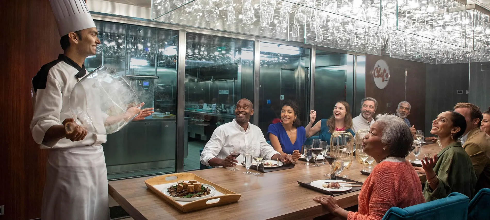 Chef presenting dish to diverse group of diners in modern restaurant kitchen