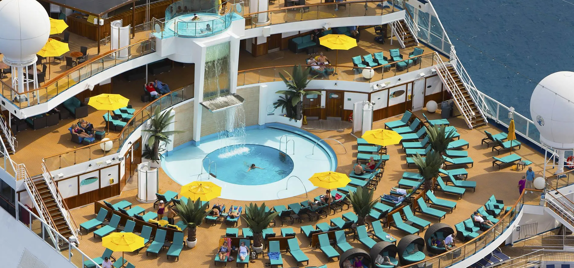 Cruise ship deck with circular pool, yellow umbrellas, and teal lounge chairs