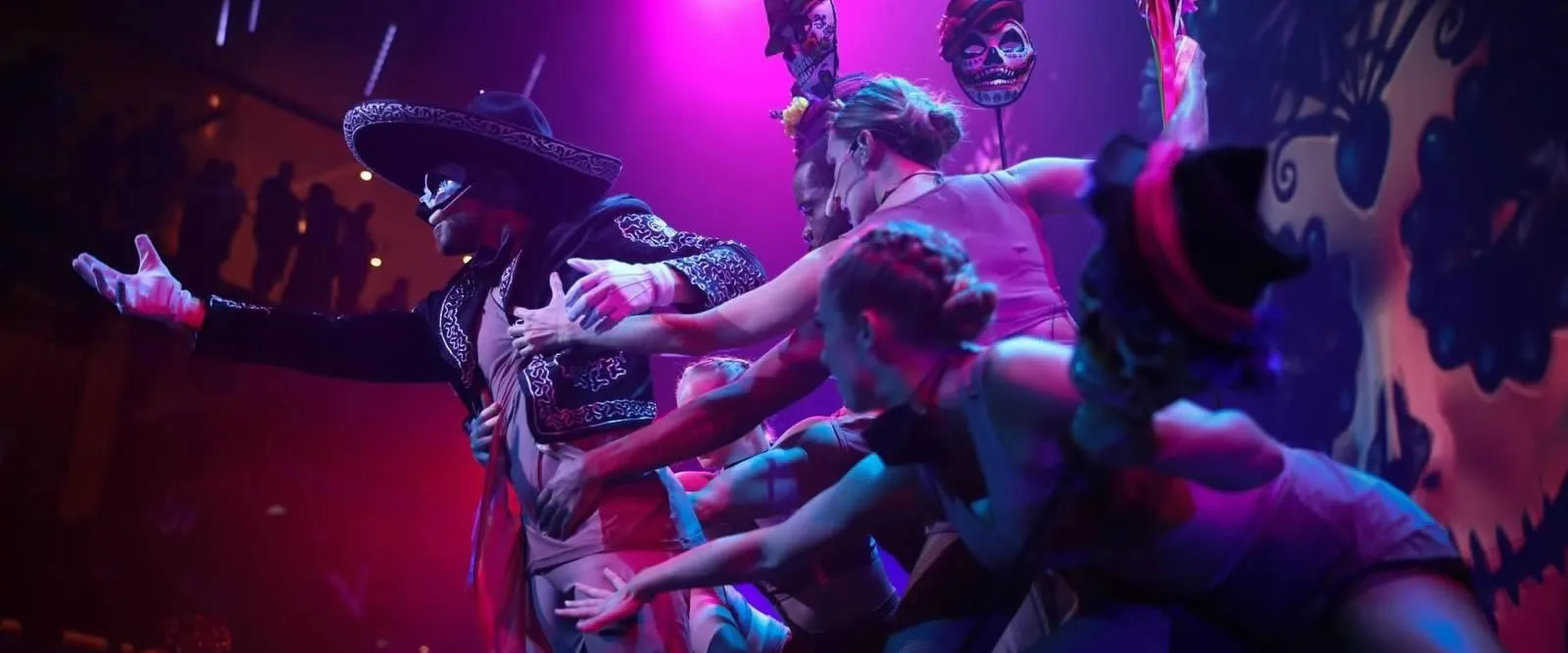 Dancers in colorful costumes perform with vibrant stage lighting and skull masks
