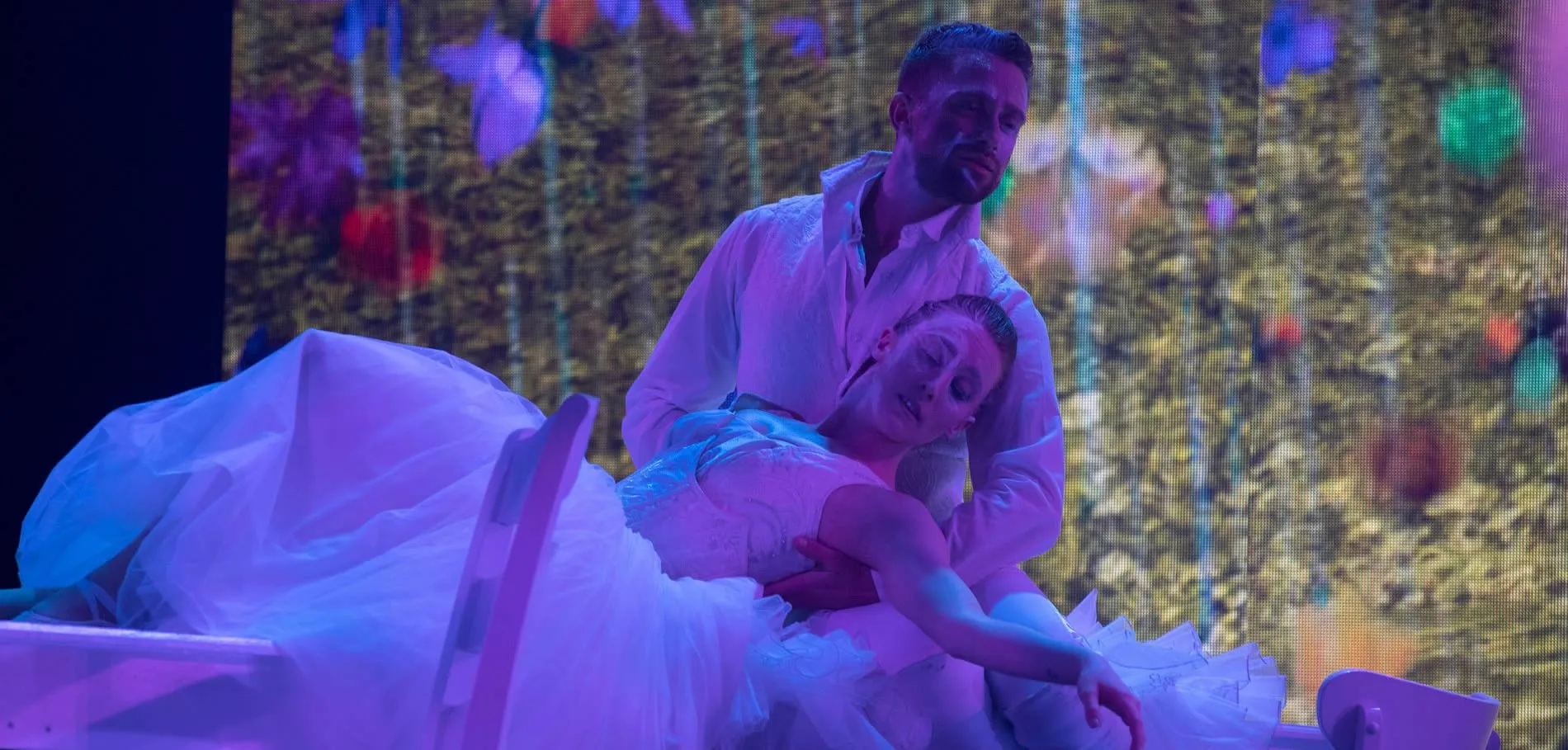 Performer in white dress held by partner on colorful theatrical stage