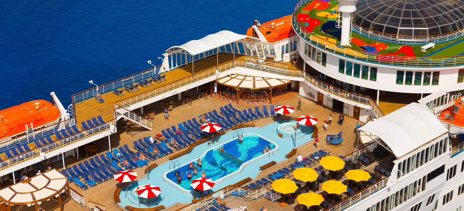 Cruise ship deck with swimming pools, blue lounge chairs, and colorful umbrellas