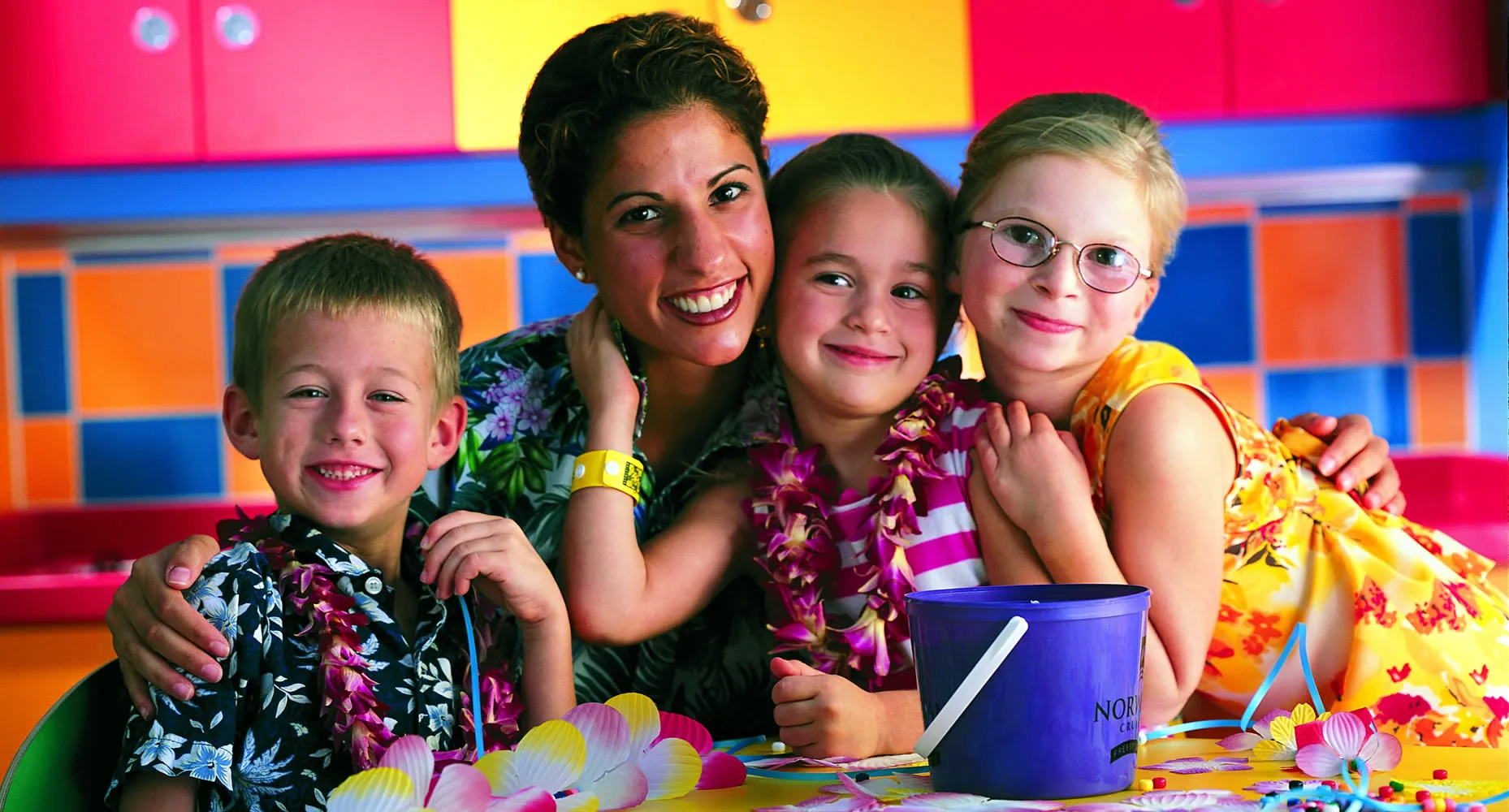 Smiling teacher with three children in colorful classroom, wearing leis