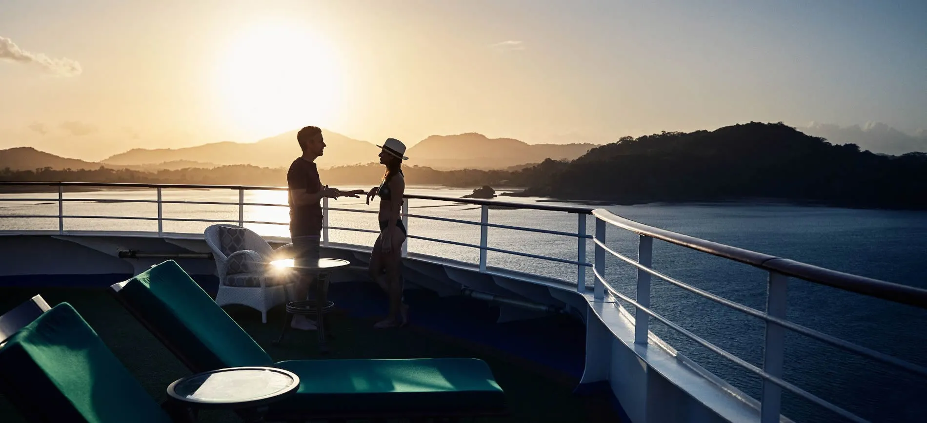 Two people on a boat deck at sunrise, with mountains and water in background