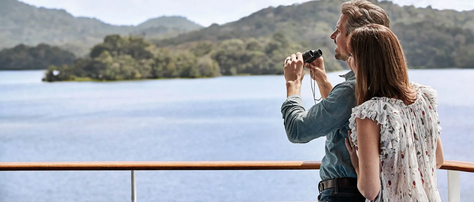 Couple using binoculars on boat deck, overlooking scenic mountain lake