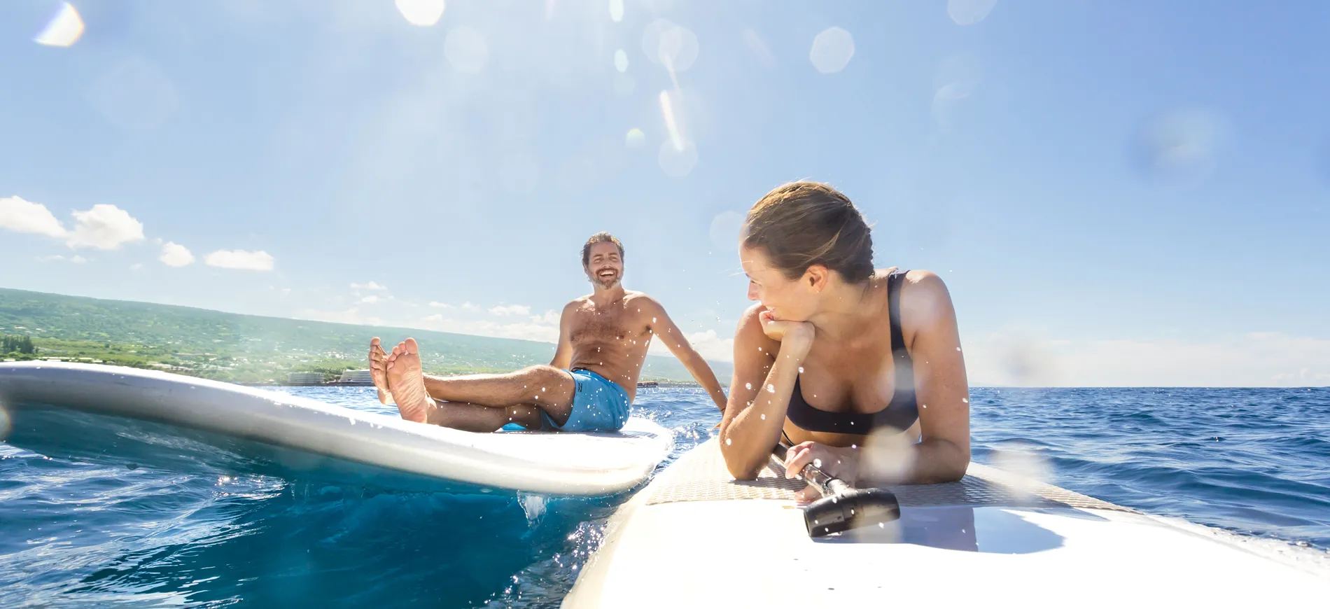 Two surfers resting on paddleboards in sunny ocean with coastal landscape