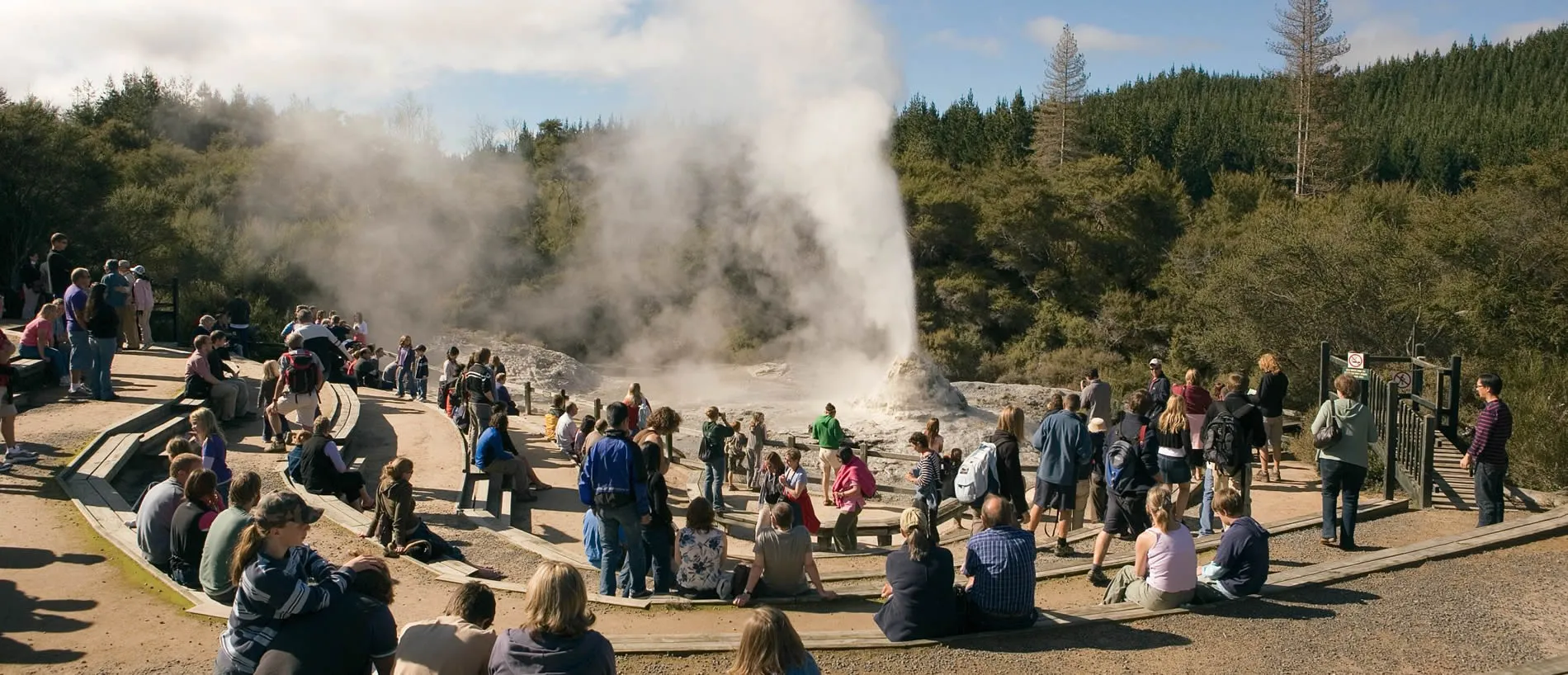 Crowded viewing platform with tourists watching steaming geyser in forest