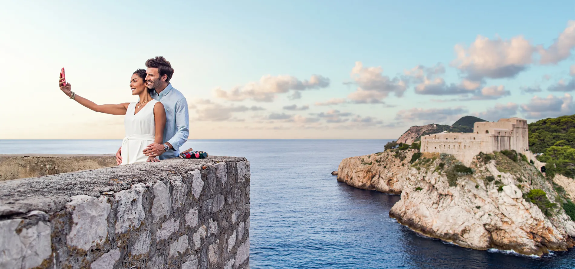 Couple taking selfie by rocky coast with historic fortress in background