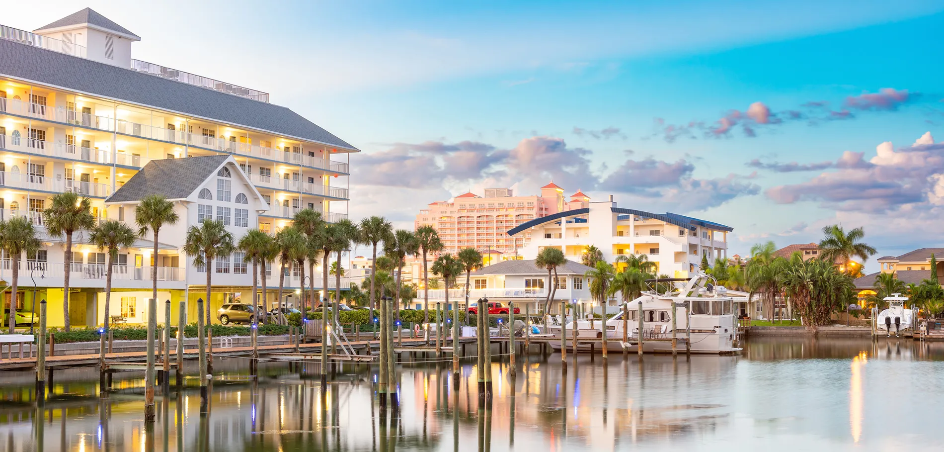 Waterfront resort with boats, palm trees, and illuminated buildings at dusk