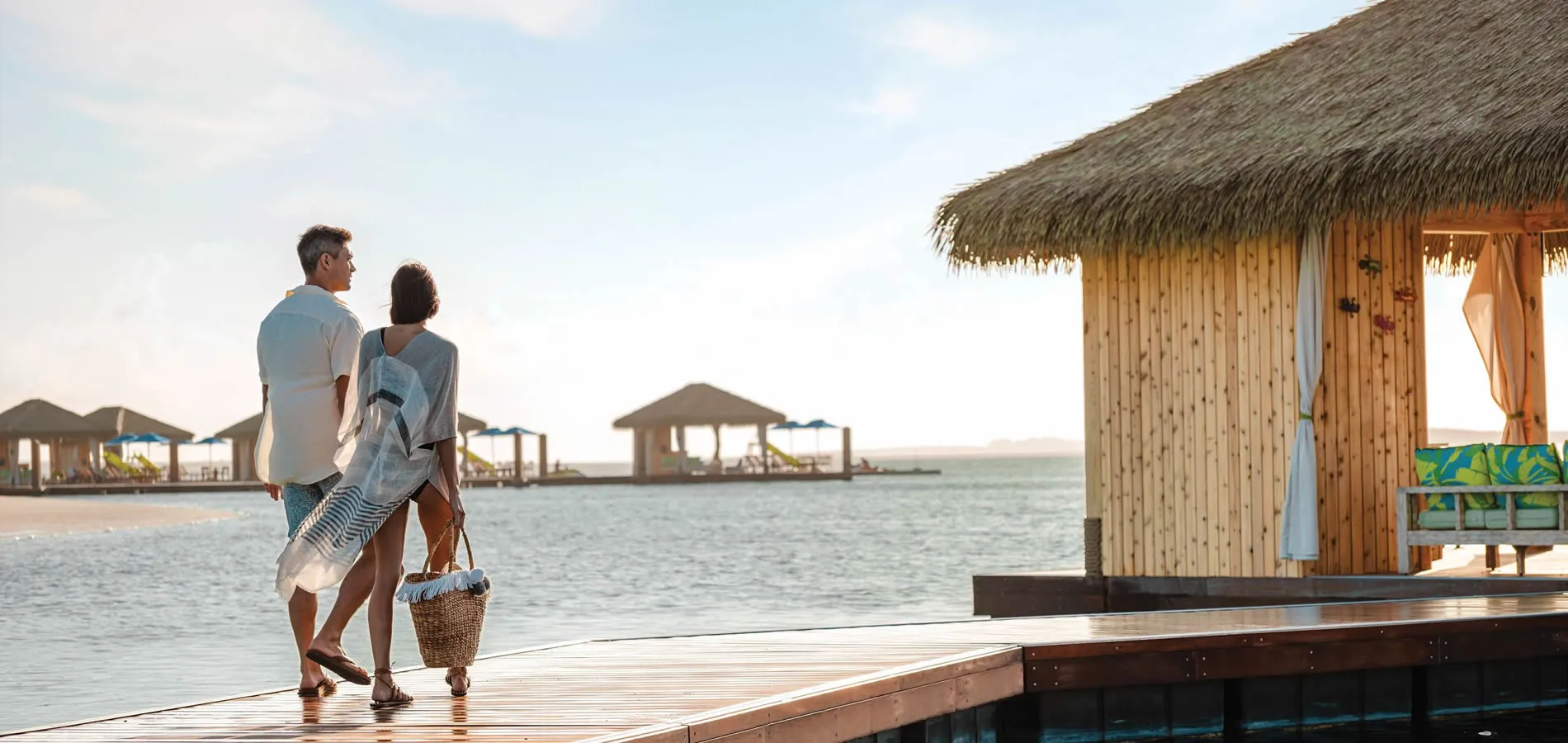 Couple walking on wooden pier near thatched beach cabanas at sunset