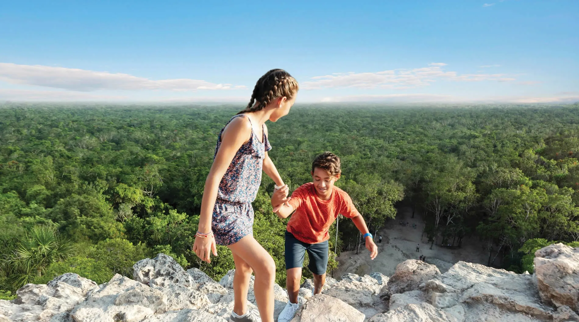 Two children hiking on rocky ledge overlooking vast green forest landscape