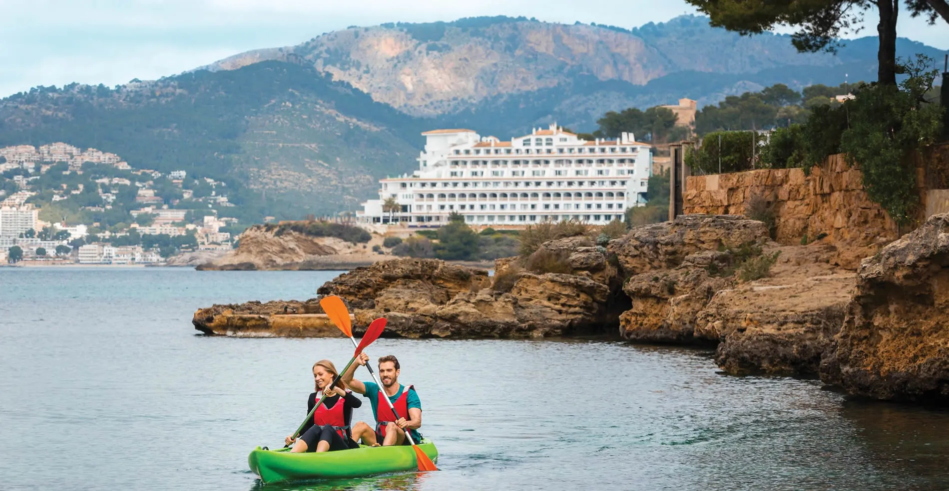 Two people kayaking near rocky coast with white hotel and mountain backdrop