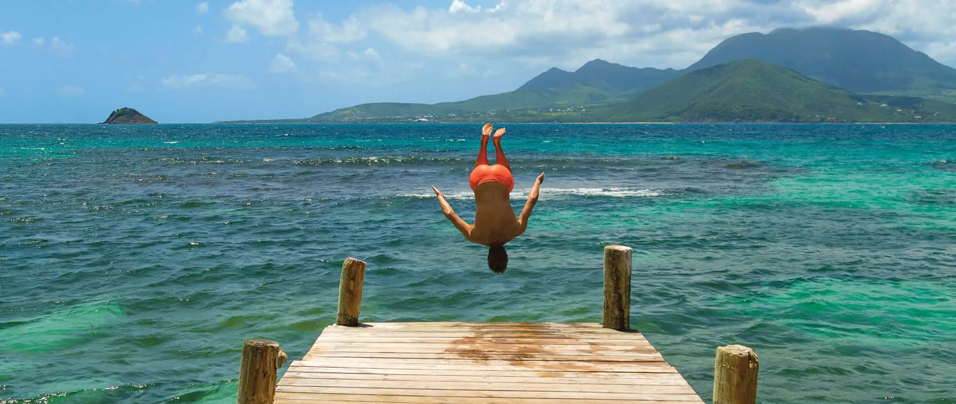 Person diving backwards into turquoise sea from wooden dock with mountains