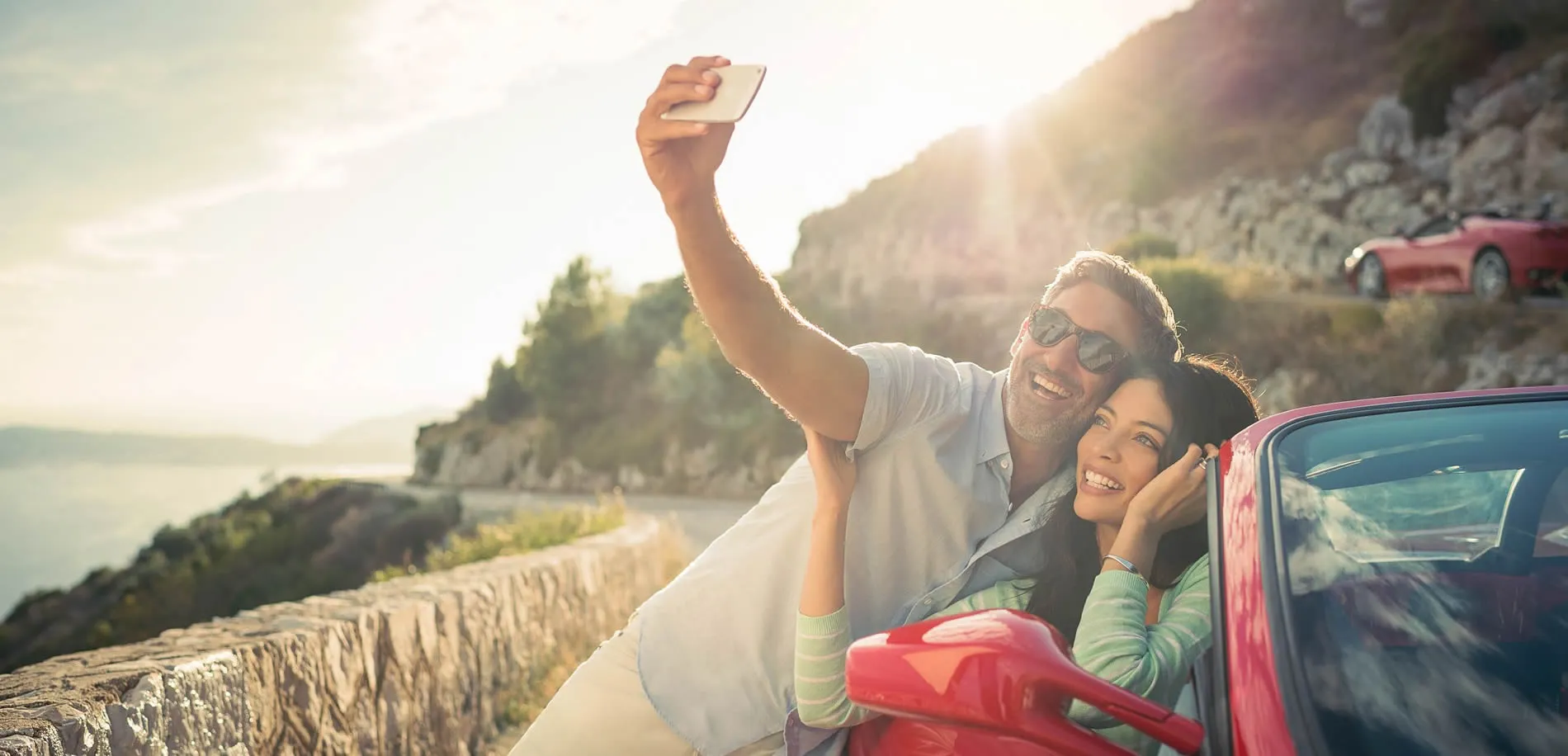 Couple taking selfie by red car on scenic coastal road at sunset