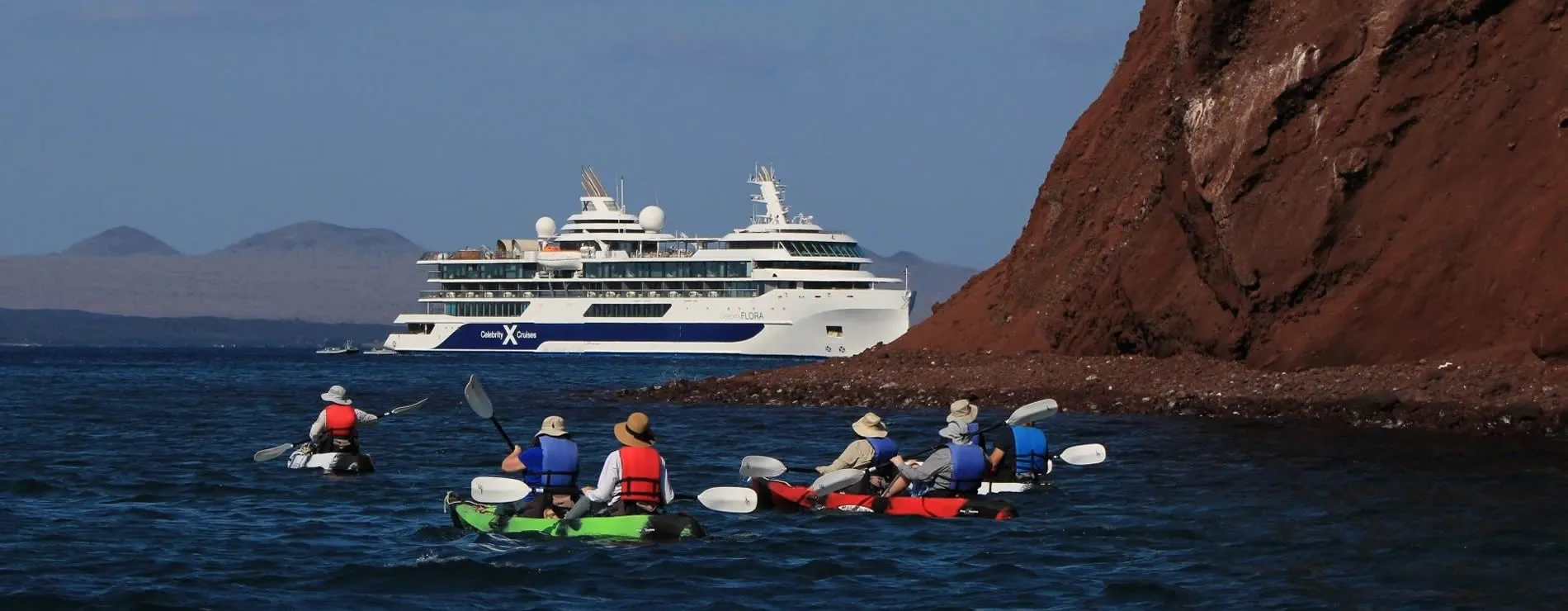 Kayakers paddle near cruise ship by red rocky coastline under blue sky