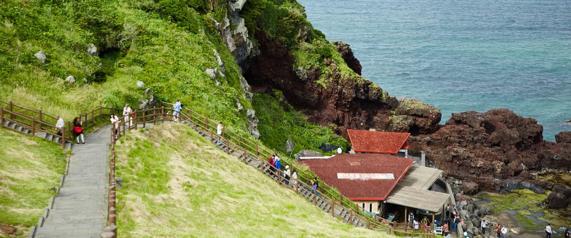 Coastal walkway with wooden path leading to red-roofed building near rocky shore