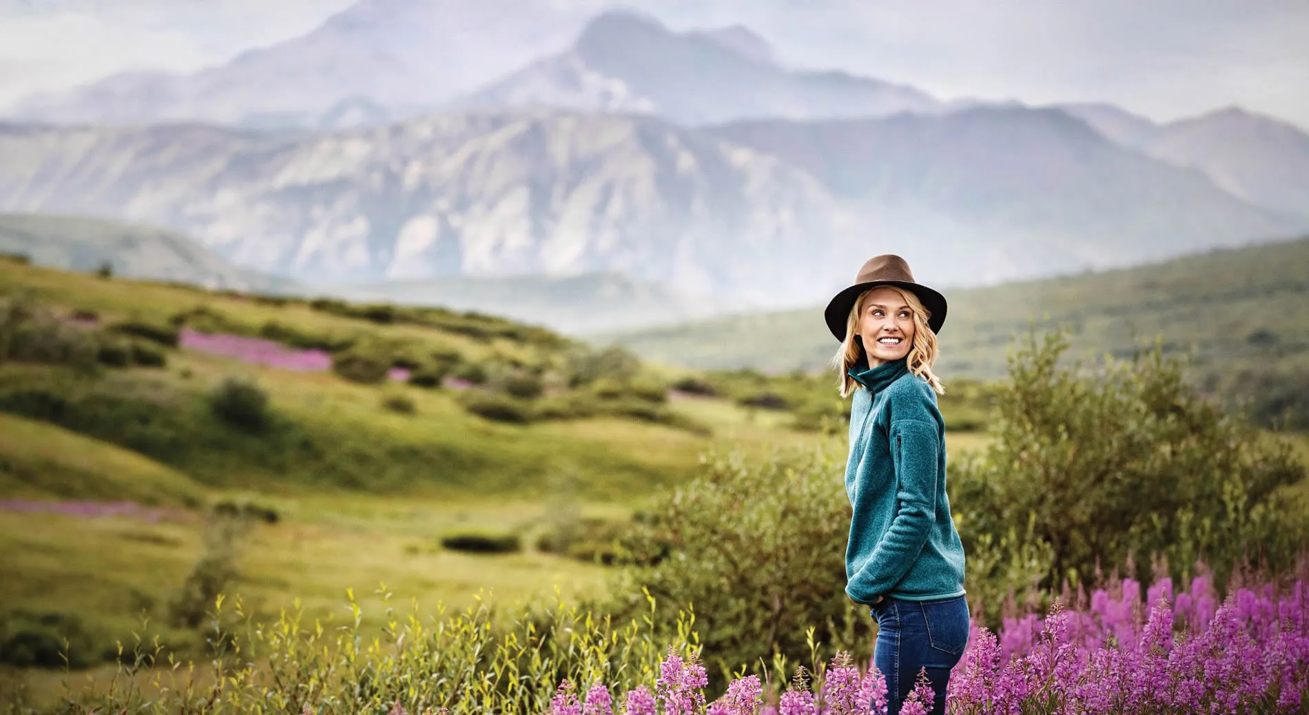 Woman in teal jacket and hat enjoying mountain landscape with wildflowers