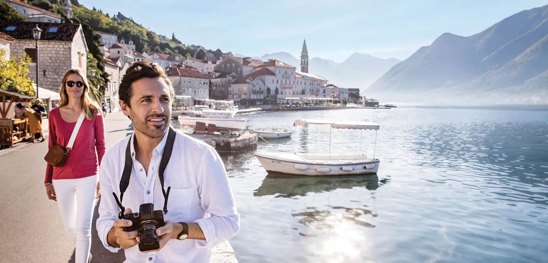 Tourists exploring picturesque coastal town with boats and mountain backdrop