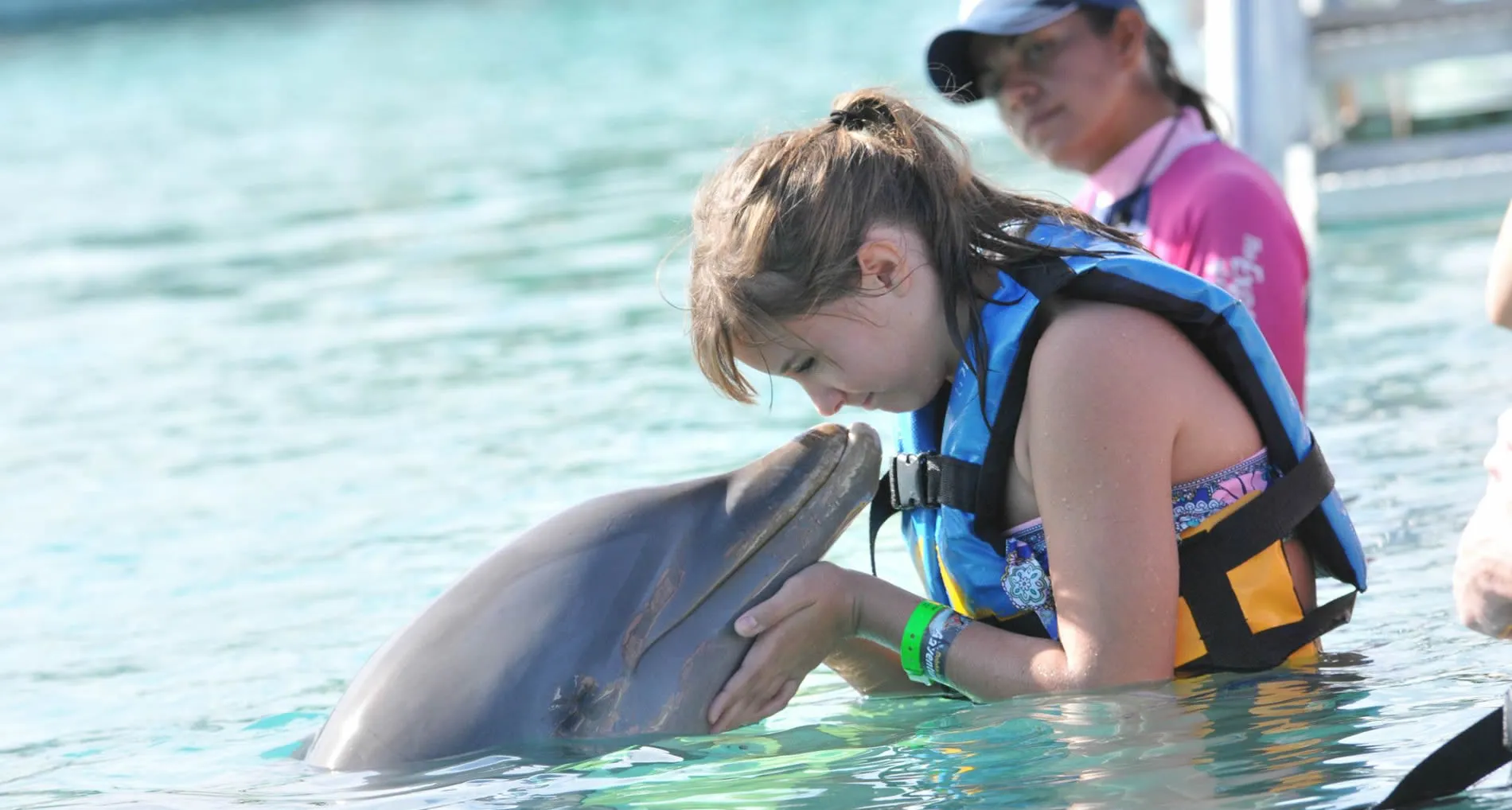 Girl in blue life vest interacts gently with dolphin in turquoise water