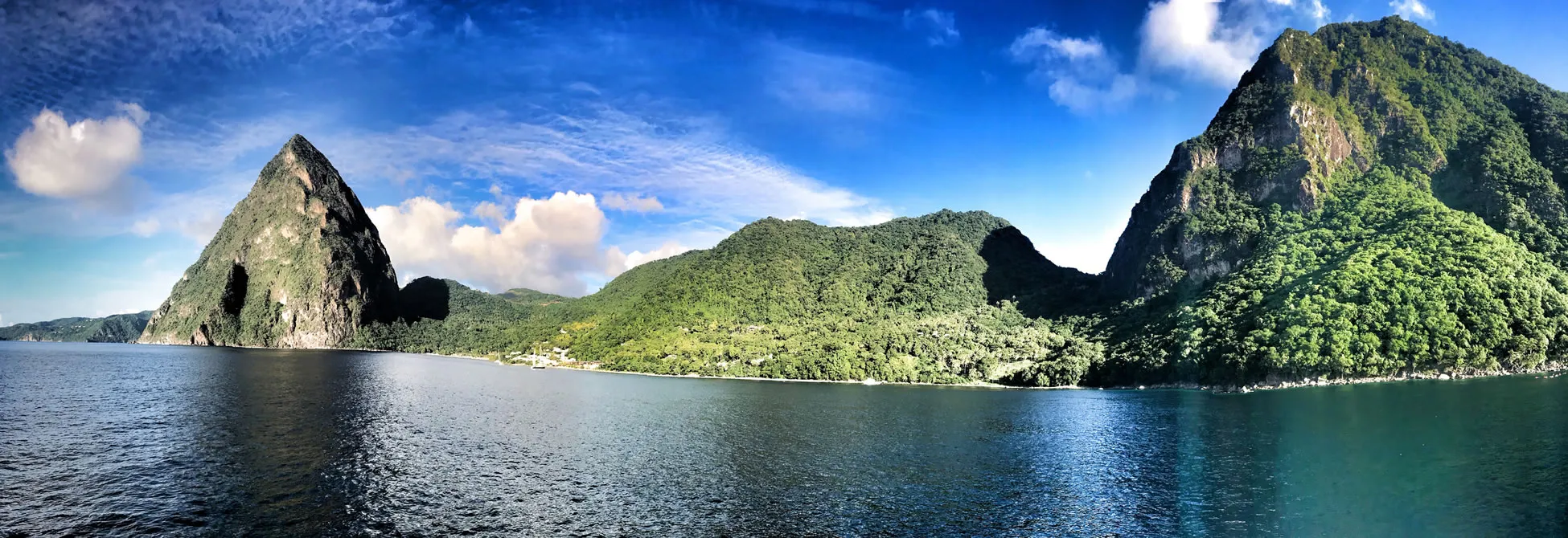 Lush green mountains rise from blue waters under bright cloudy sky