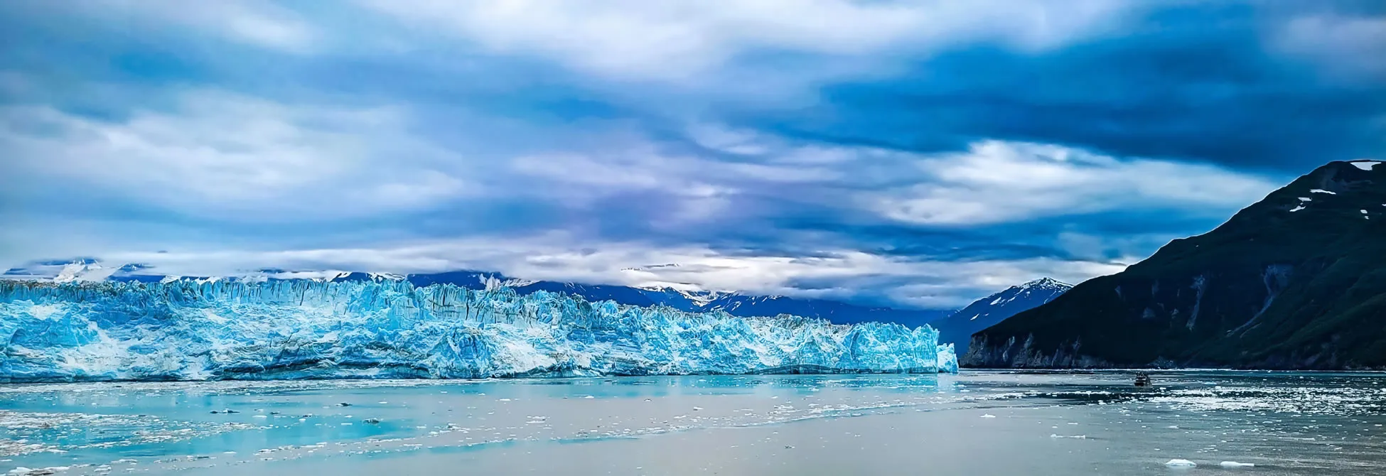 Massive blue glacier meets turquoise water under dramatic cloudy sky