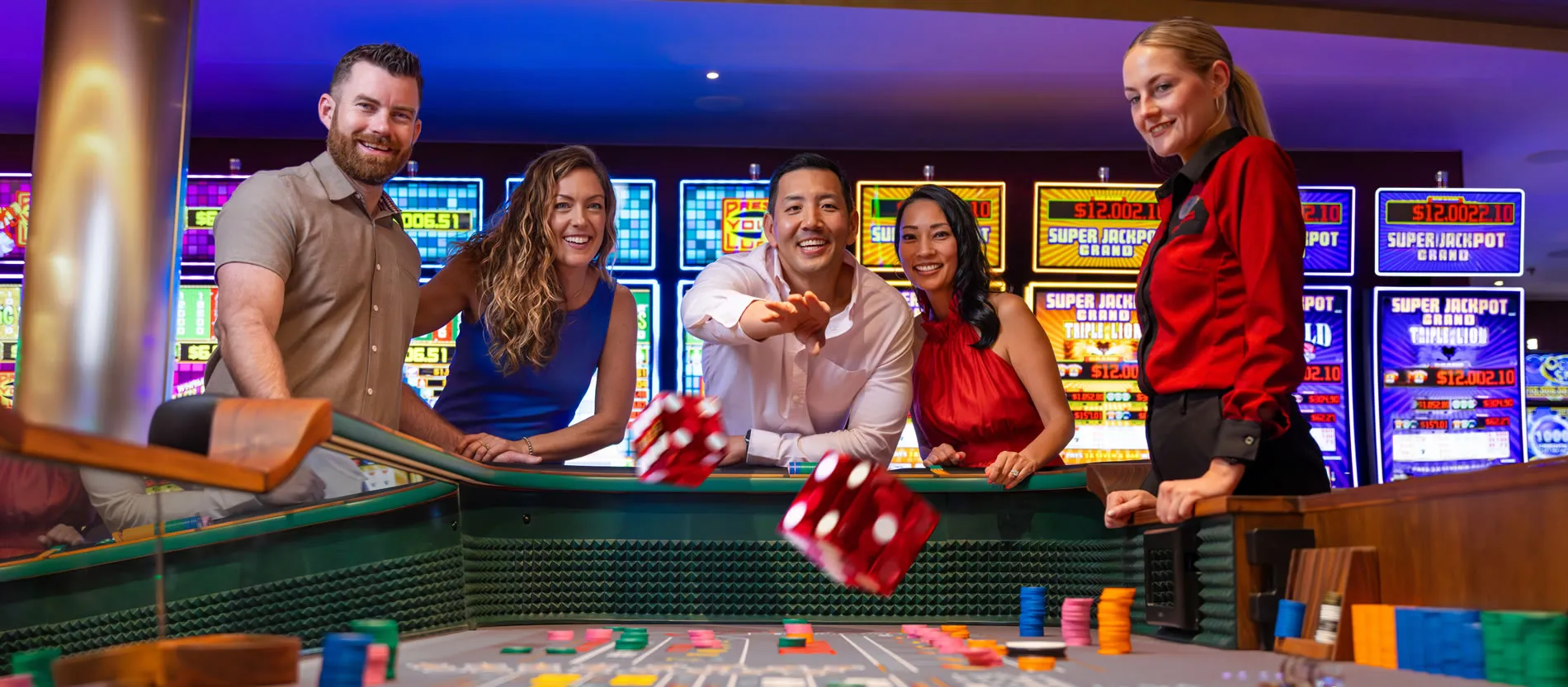 Casino group throws dice at craps table with slot machines in background