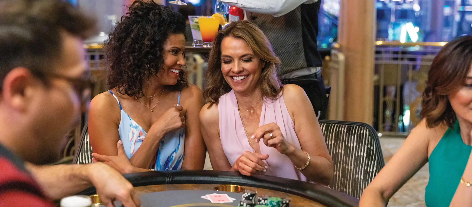Two women laughing together at a casino gambling table with drinks