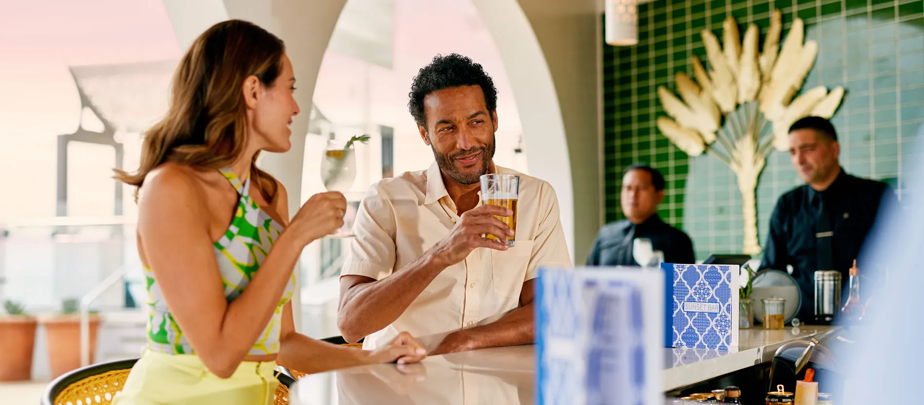 Two people enjoying drinks at a bar with decorative palm leaf backdrop