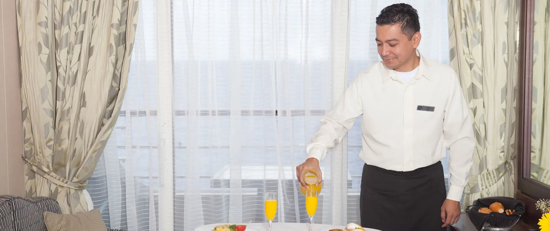 Waiter in white uniform pouring orange juice by window with curtains