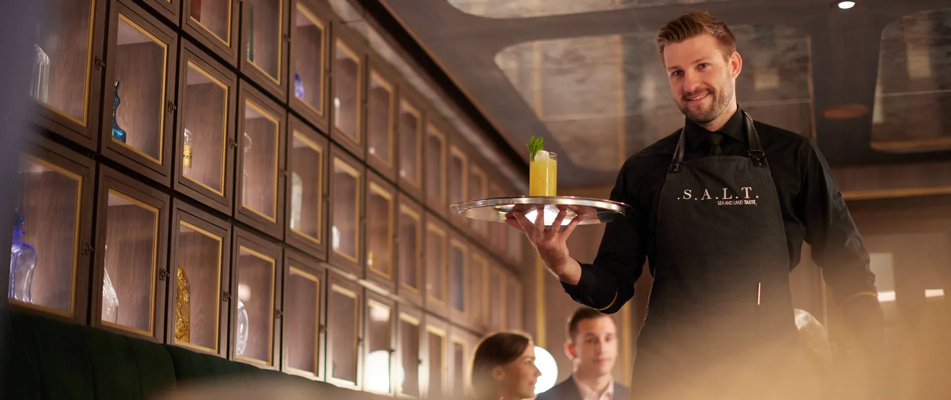 Bartender in black S.A.L.T. apron holding yellow cocktail on silver tray