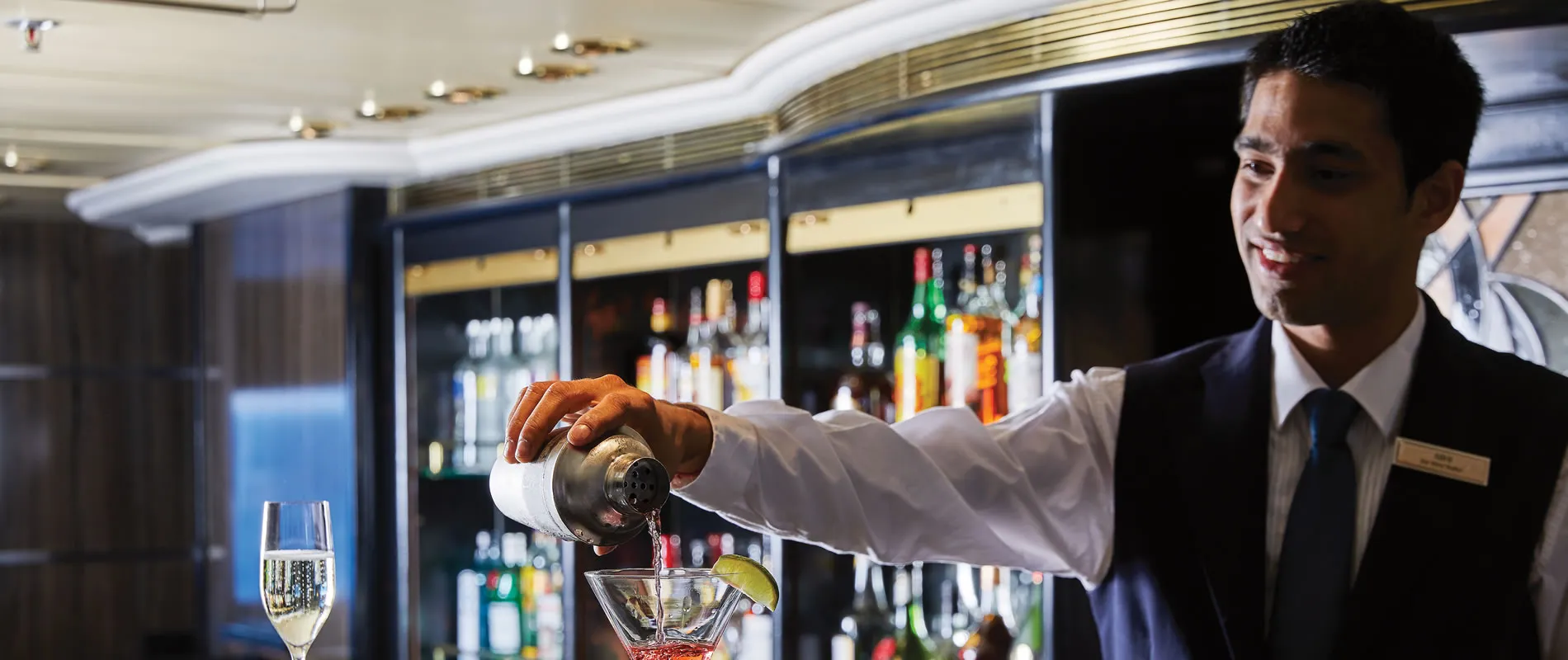 Bartender in suit pouring cocktail at bar with liquor bottles behind