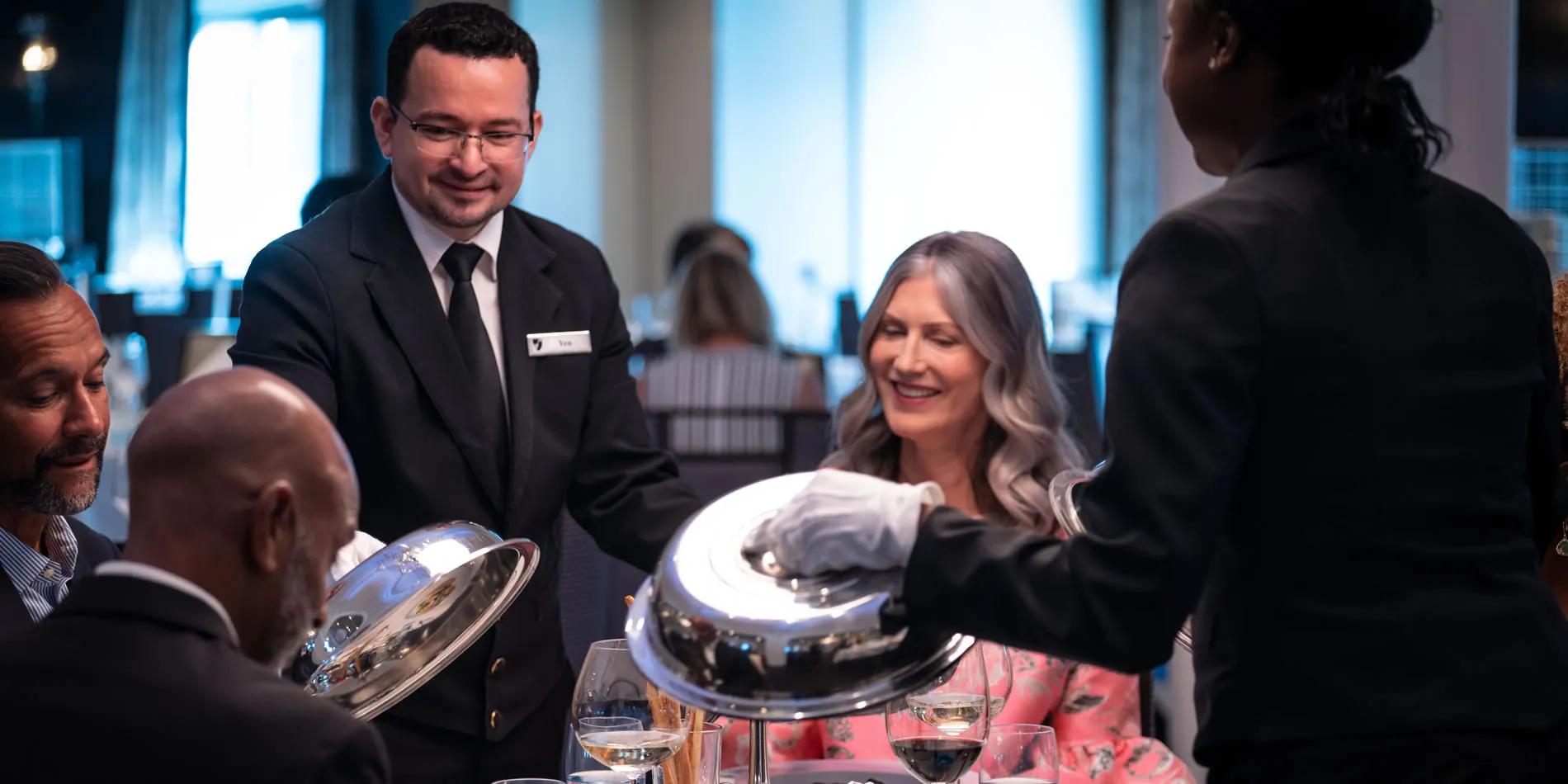 Restaurant staff serving guests with silver covered dishes and wine