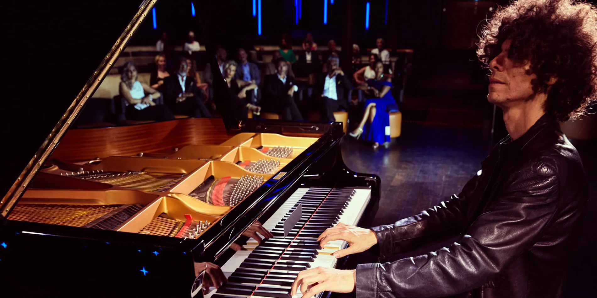 Pianist with curly hair performs on grand piano with audience in background