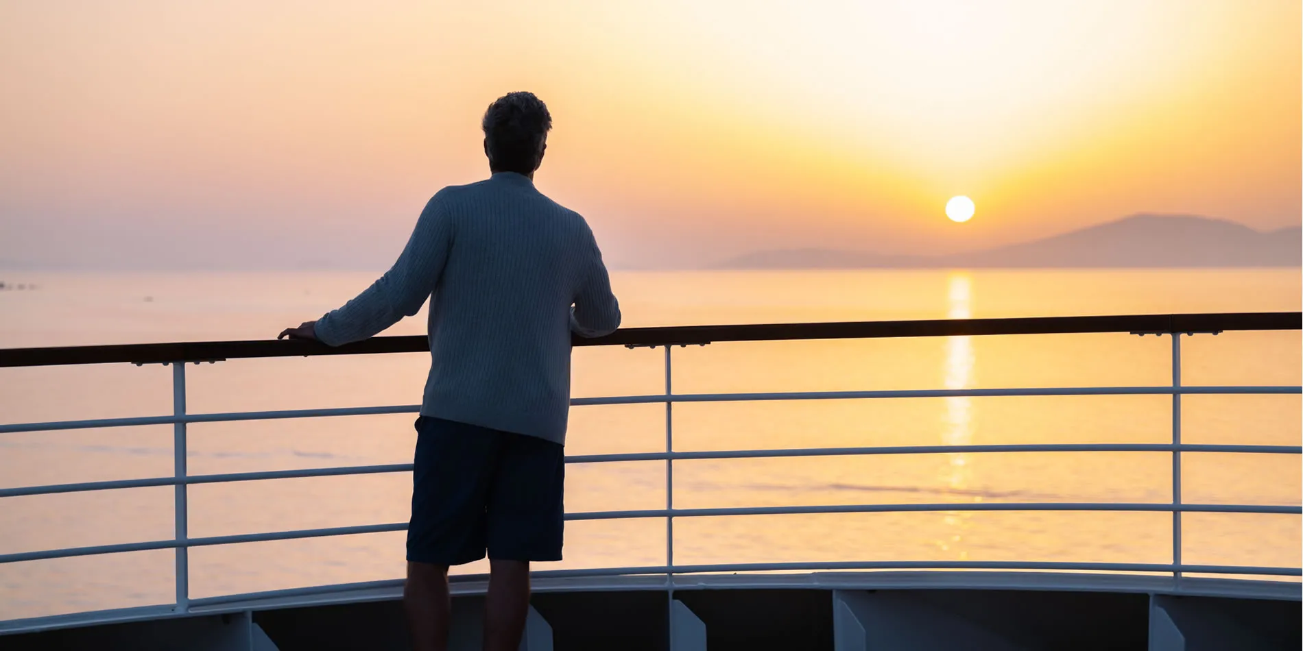 Person on boat deck watching sunrise over misty mountains and calm water