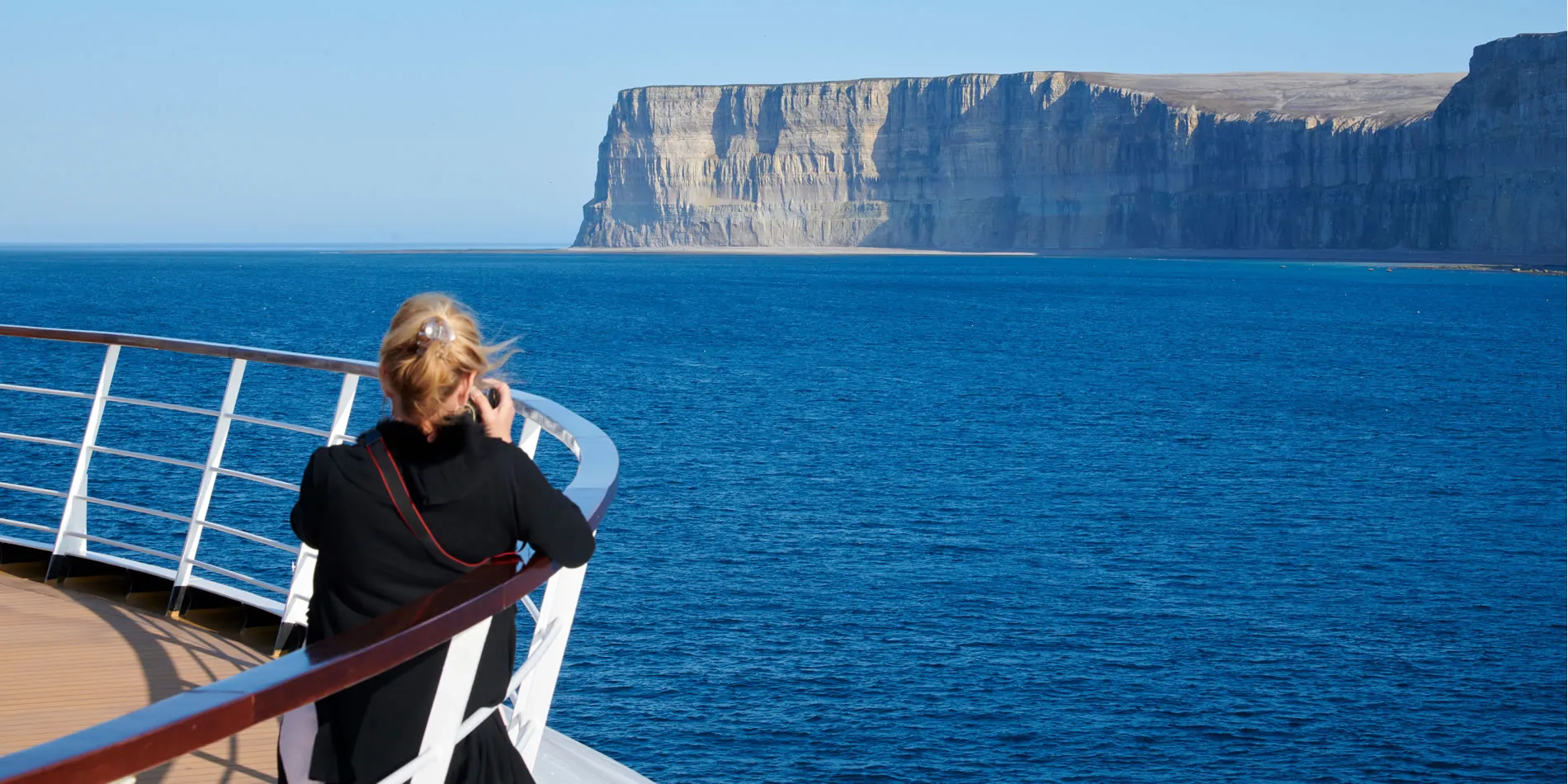 Person on cruise ship deck viewing dramatic coastal cliffs and blue ocean